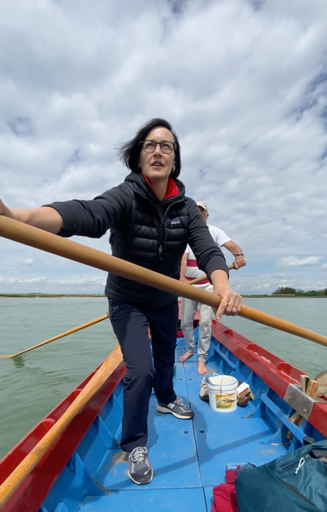 Artist Melissa McGill standing in a gondola with boats large oar in hand leaning forward, on a waterway of Turin.