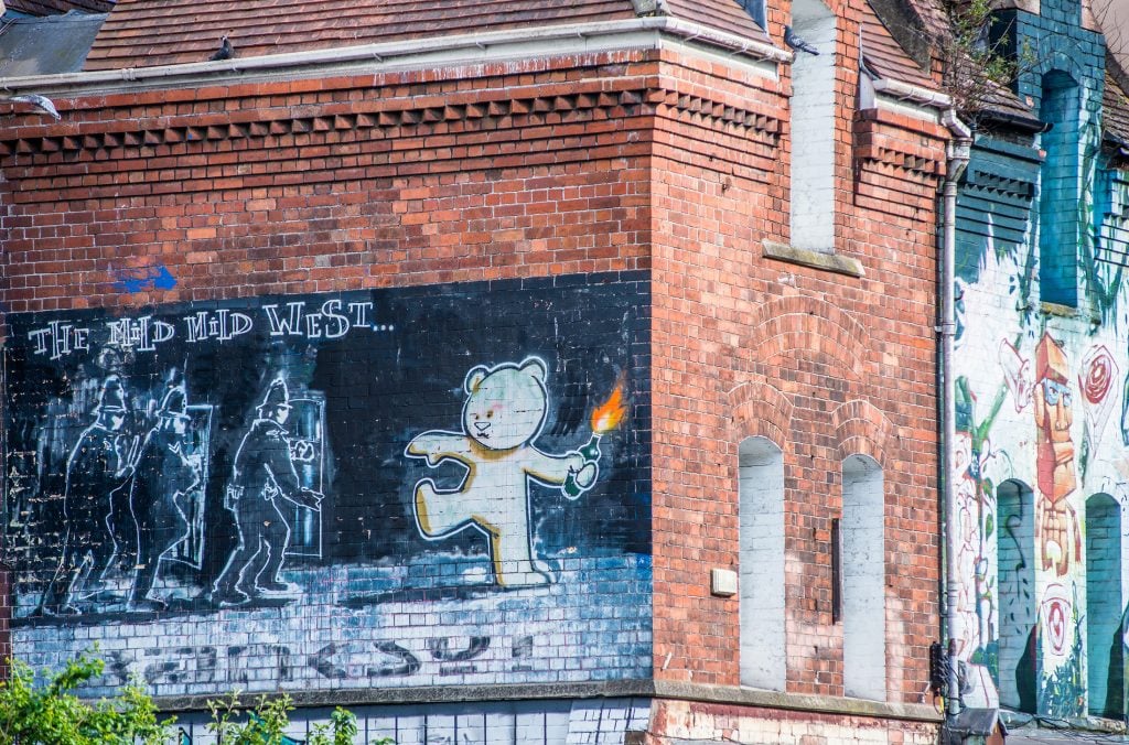 A black and white mural by Banksy depicting a teddy bear hurling a molotov cocktail at a group of police offers, painted on a brick wall