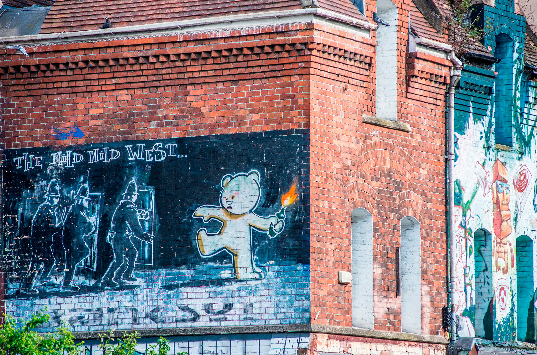 A black and white mural by Banksy depicting a teddy bear hurling a molotov cocktail at a group of police offers, painted on a brick wall