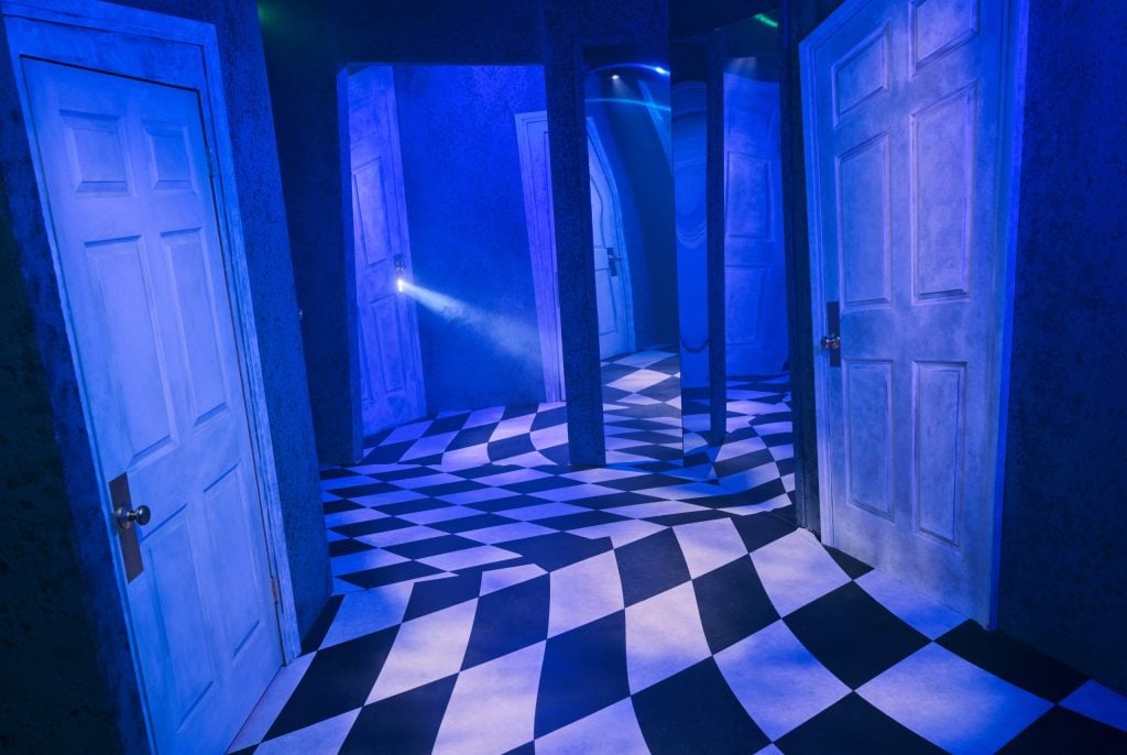 a warped hallway with odd angles with a tiled black and white floor and large white doors cast in a radiant blue light