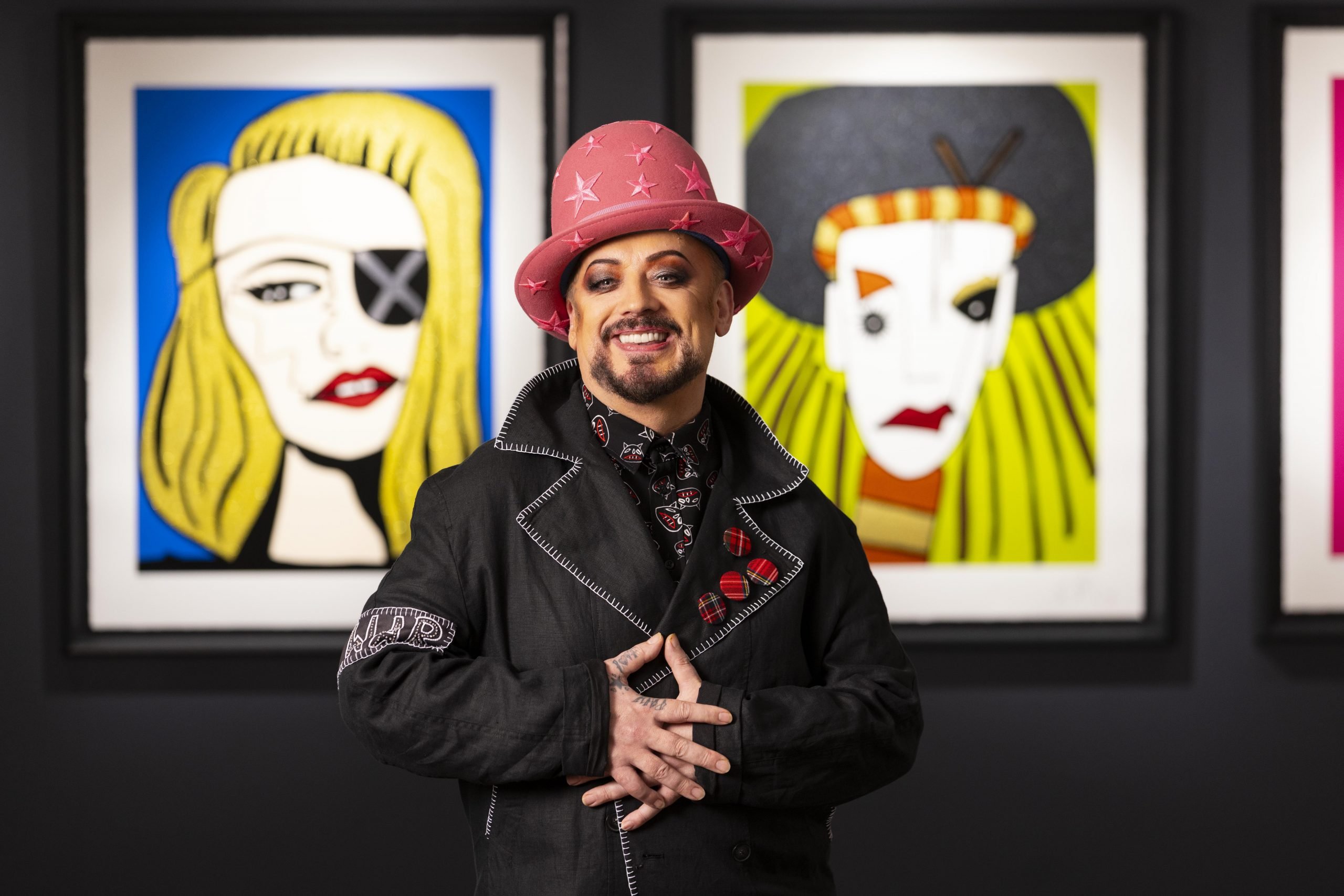 Singer Boy George posing in a pink hat in front of two of his paintings