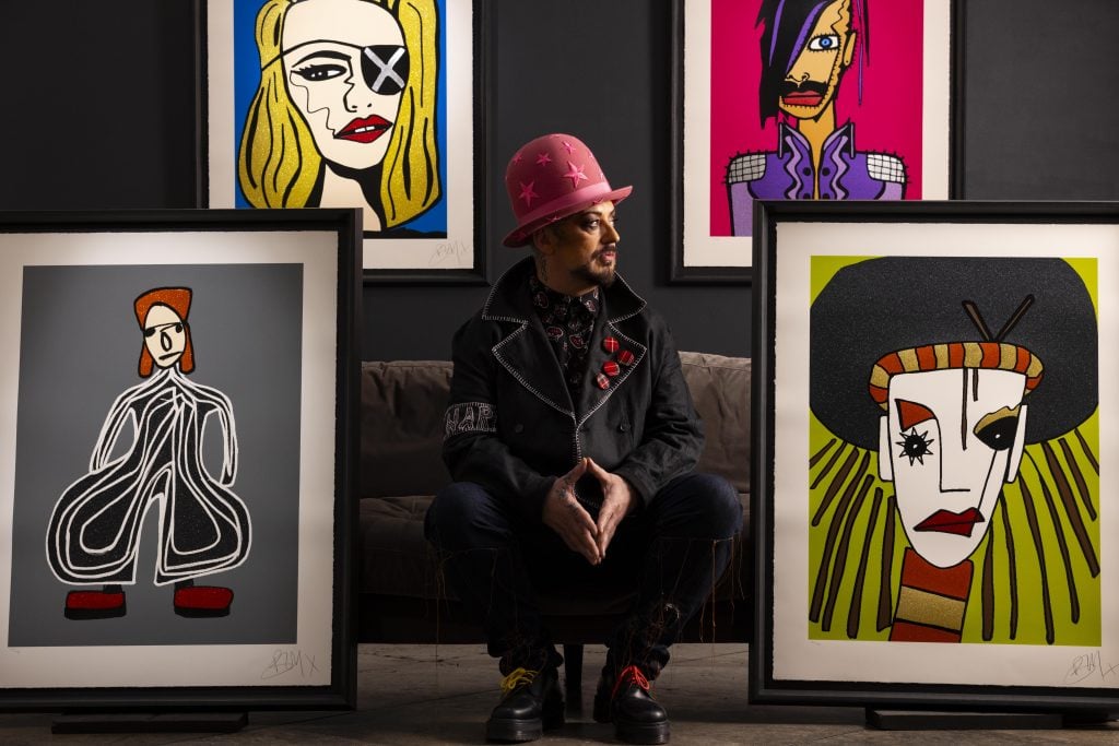 Boy George sitting on a sofa surrounded by four of his paintings