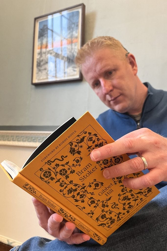 A man holding open a book with a cover that reads "Gibbet Hill" by Bram Stoker