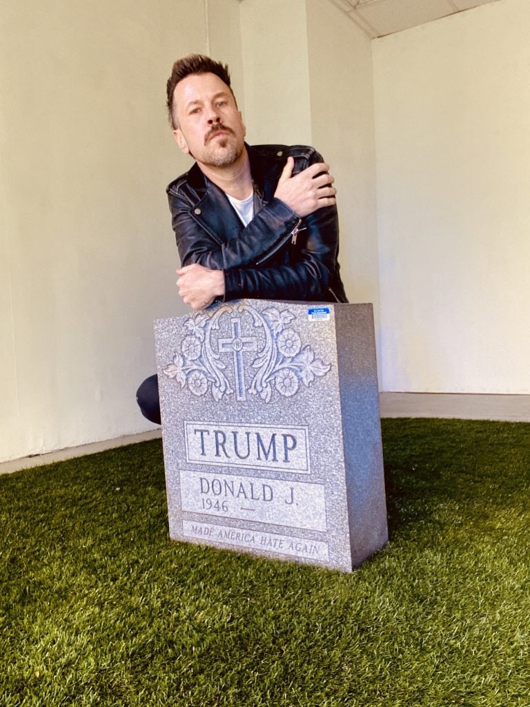 Brian Andrew Whiteley with Legacy Stone, the Donald Trump tombstone that he left as a guerrilla artwork in Central Park on Easter Sunday, March 2016. The artist, a young white man in a black leather jacket, squats behind a traditional granite gravestone with ornamental carving of a cross and an inscription for Donald J. Trump reading 