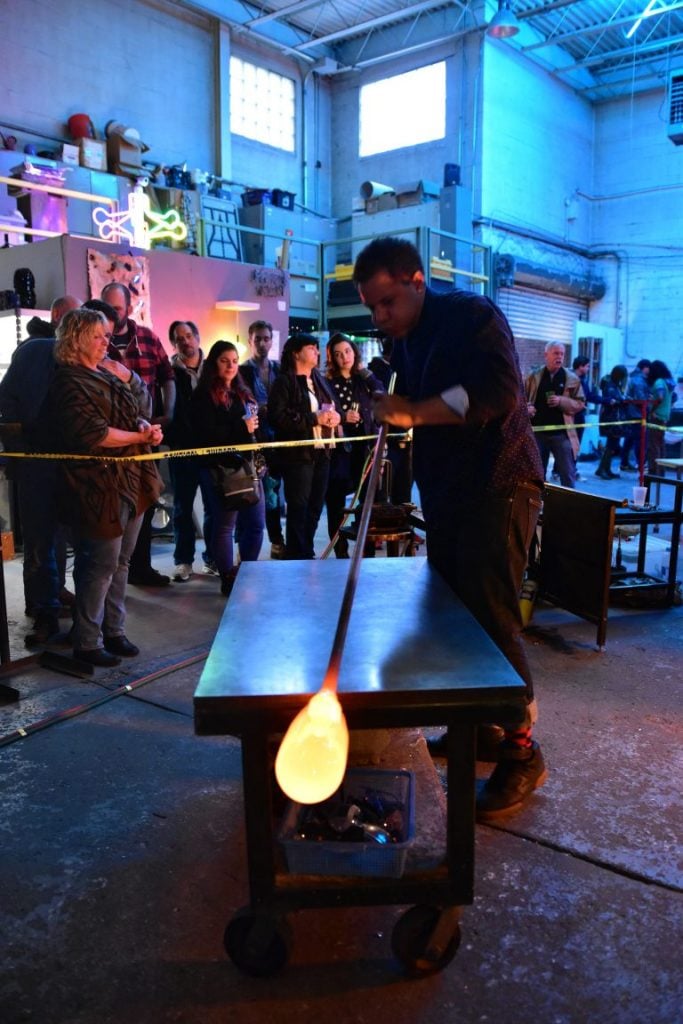 A glass blowing workshop at Brooklyn Glass. A crowd of onlookers stand behind caution tape as a man rolls hot glass at a workstation.