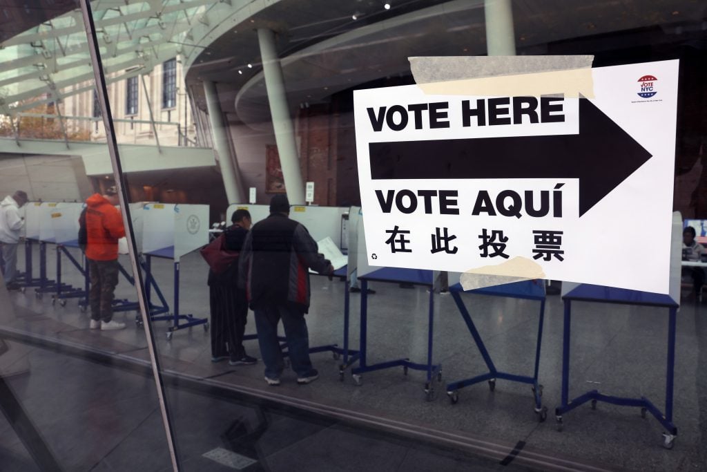 People voting at the Brooklyn Museum in 2023 in New York City. The voting booths in the museum lobby are seen through a glass window with a sign reading "vote here" in English and Spanish.