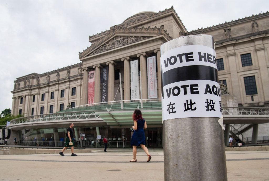 A sign reading "VOTE HERE" is stuck on a pillar in the foreground, with the Brooklyn Museum in the background