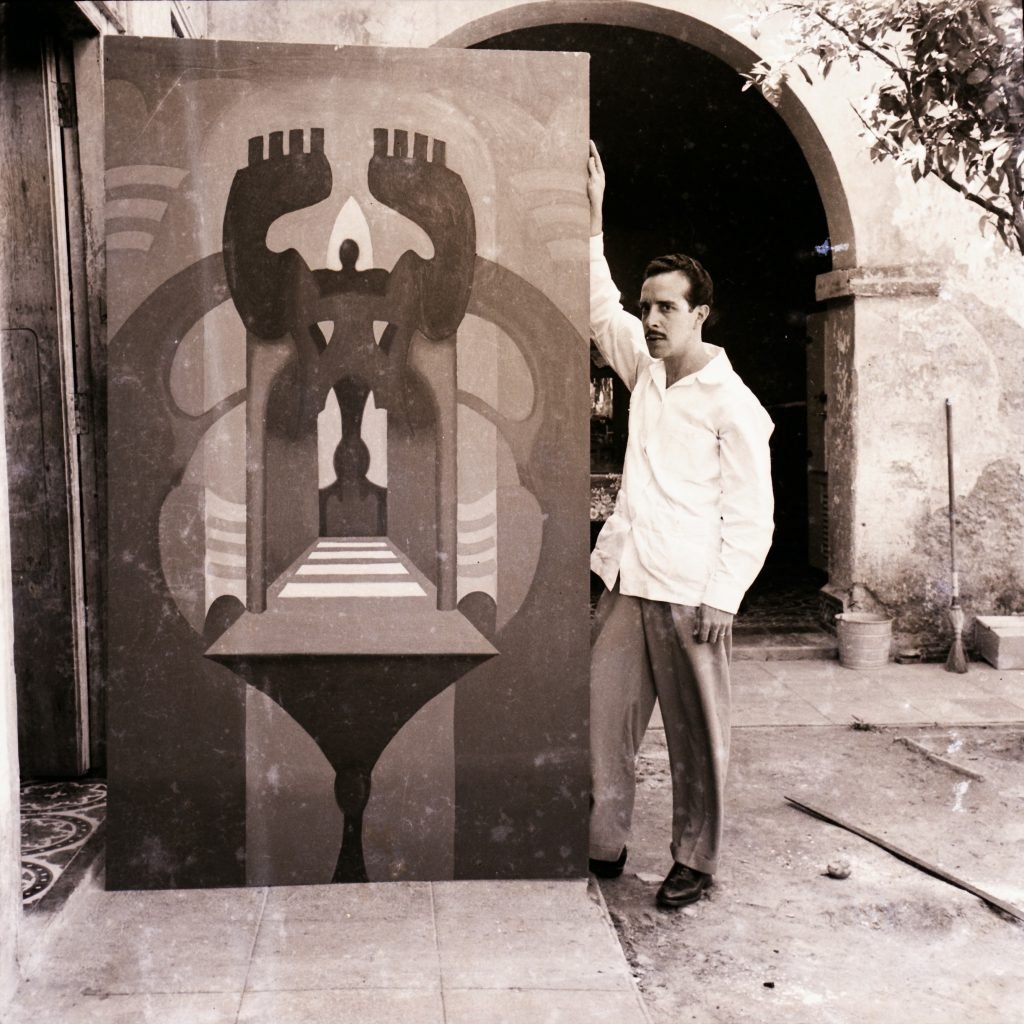 A black and white photo of a young Rafael Soriano in Cuba, standing in a courtyard behind an archway with a monumental abstract painting of geometric forms.