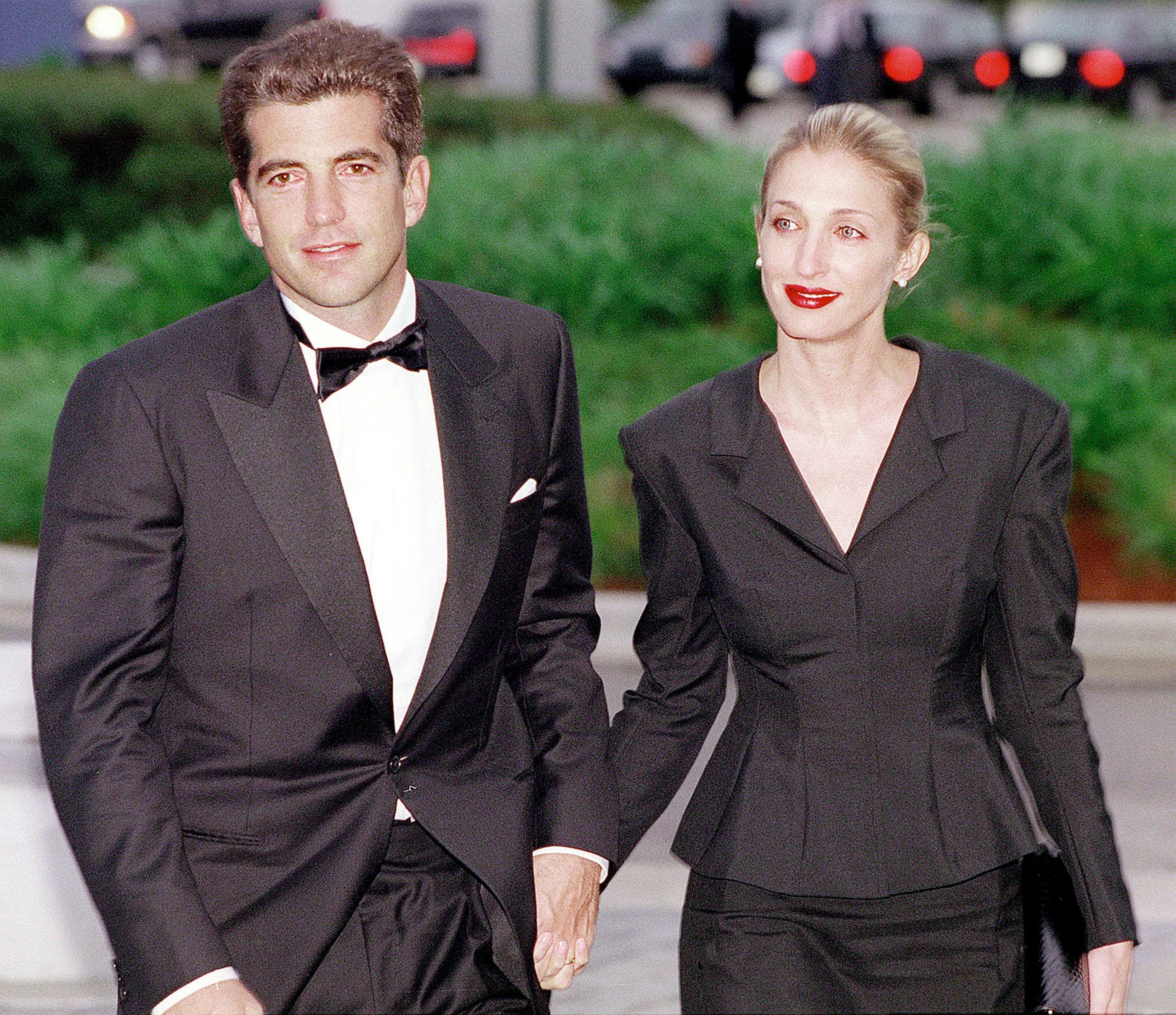 John F. Kennedy Jr. in a tuxedo posing alongside Carolyn Bessette-Kennedy in a black dress suit