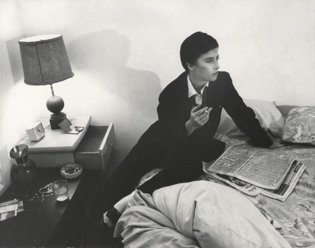 Black and white photograph of the photographer Cindy Sherman dressed as a man in a black suit smoking and leaning across a bed in a bedroom, featured in Artnet Auctions' Important Photographs sale.