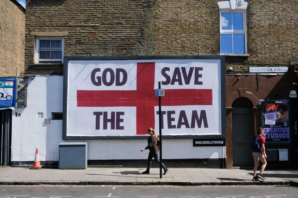 A person walking past a large billboard with a large red cross in its center and the words 'God Save the Team'