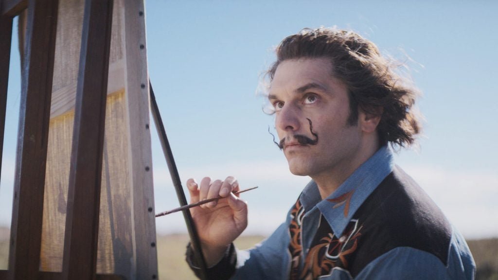 In a still from the film Daaaaaalí, a man with chin-length hair and a wiry mustache holding a paint brush against a canvas on an easel