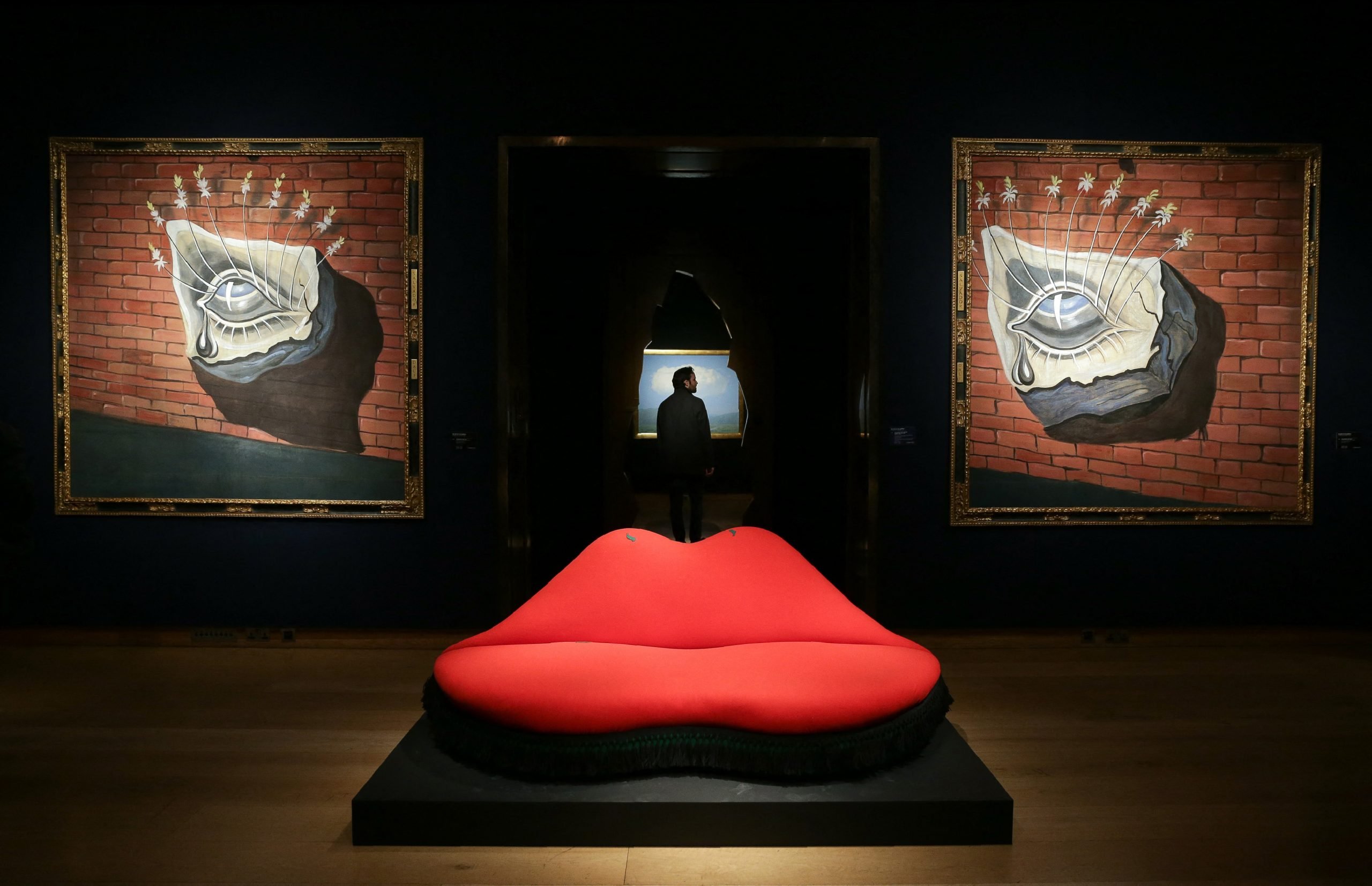 A sofa shaped after lips on display in a gallery in front of two paintings of eyes