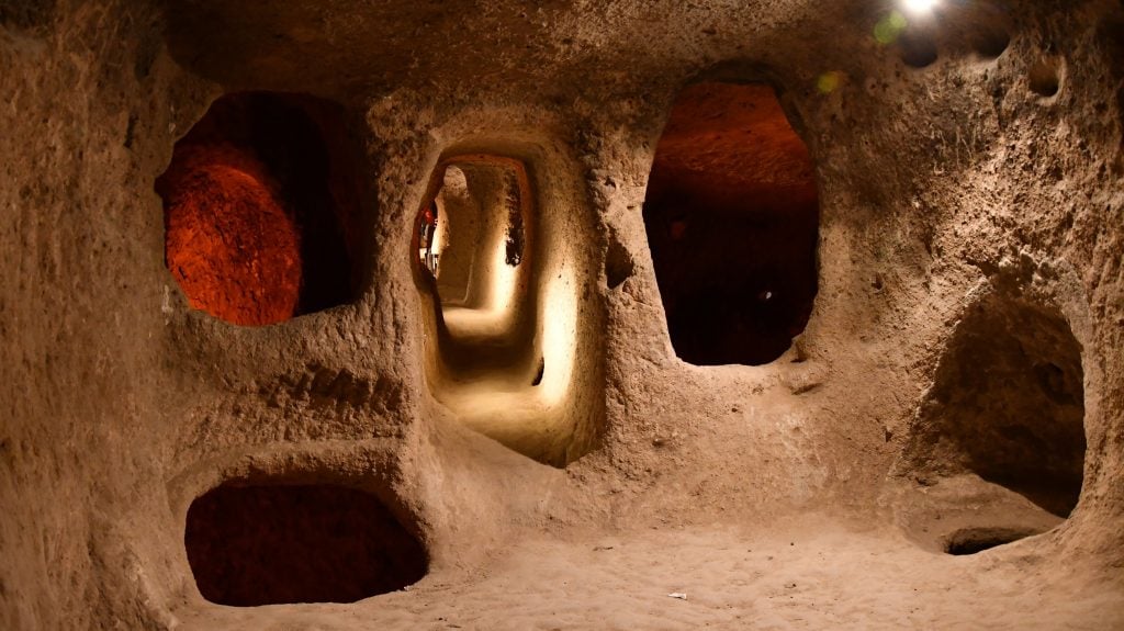 Narrow underground passageways connecting small rooms in the Derinkuyu cave system. The stone walls are naturally worn, with some areas illuminated by soft light.