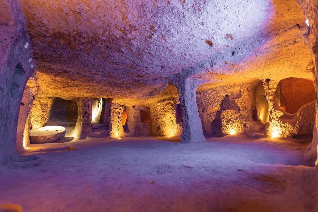 A spacious, illuminated underground chamber with arched doorways, thick support columns, and a smooth dirt floor, showing the extensive cave network of Derinkuyu in Cappadocia, Turkey.