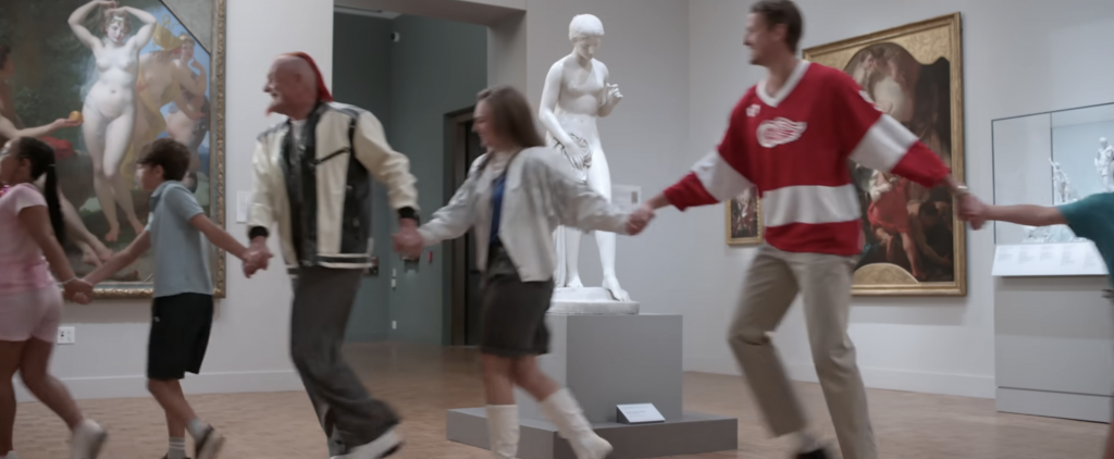 A still from "Darren McCarty's Day Off" from filming at the Detroit Institute of Arts. Three adults are hand in hand with school children passing in front of a classical sculpture.