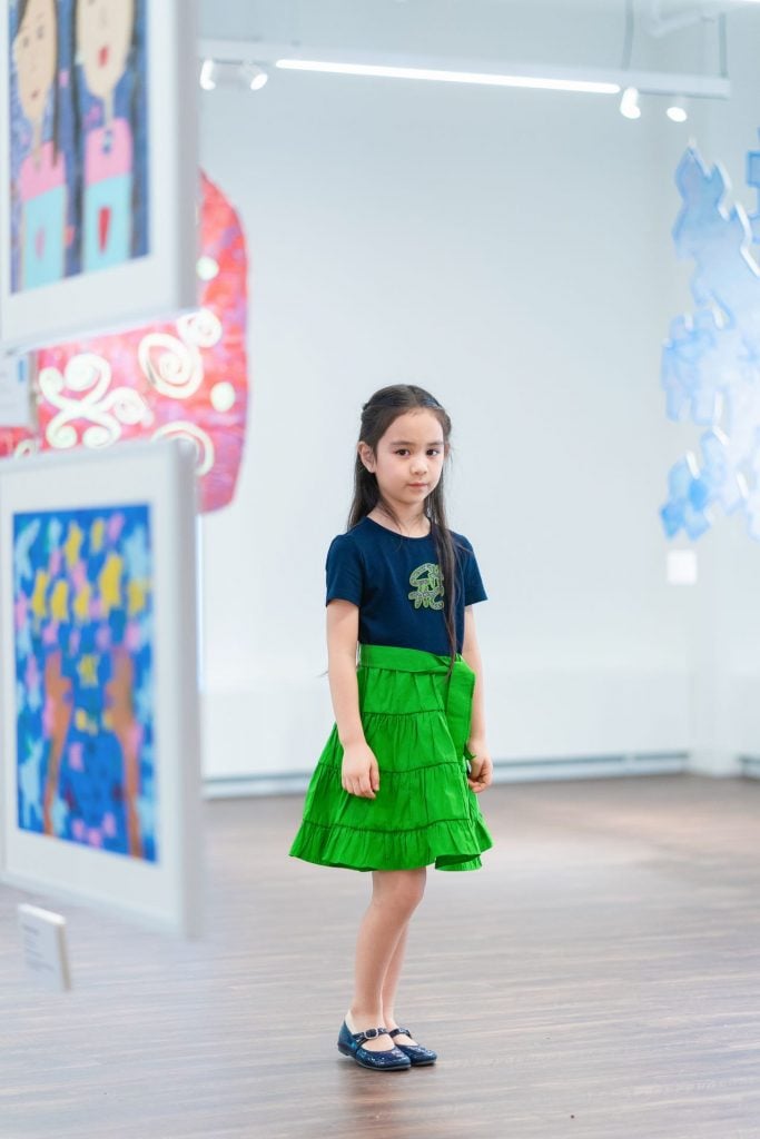 Photo of 6 year old artist Elaine Fan wearing a green skirt and black Mary Jane shoes standing in the gallery space of the China Institute surrounded by hanging paintings.