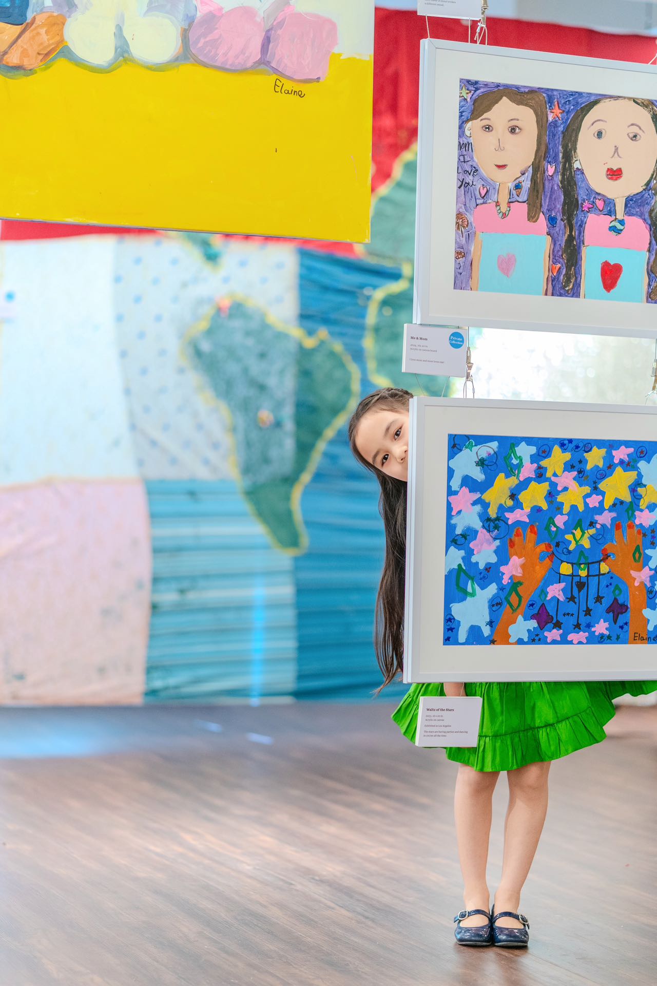 Photo of 6 year old artist Elaine Fan wearing a green skirt and black Mary Jane shoes peaking out from behind two of her paintings hanging at the China Institute.