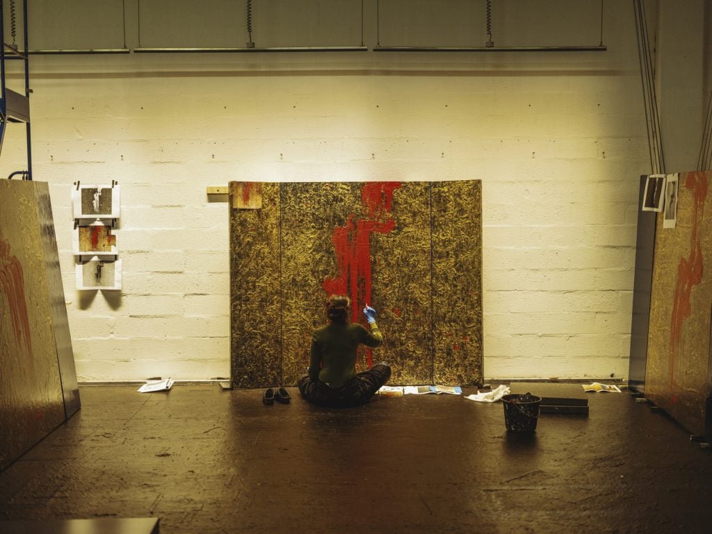 Artist Fabienne Verdier sitting cross legged on the ground with her back to the camera painting a large-scale, gold leaf canvas with swathes of vibrant red, all situated inside the artist's concrete studio.