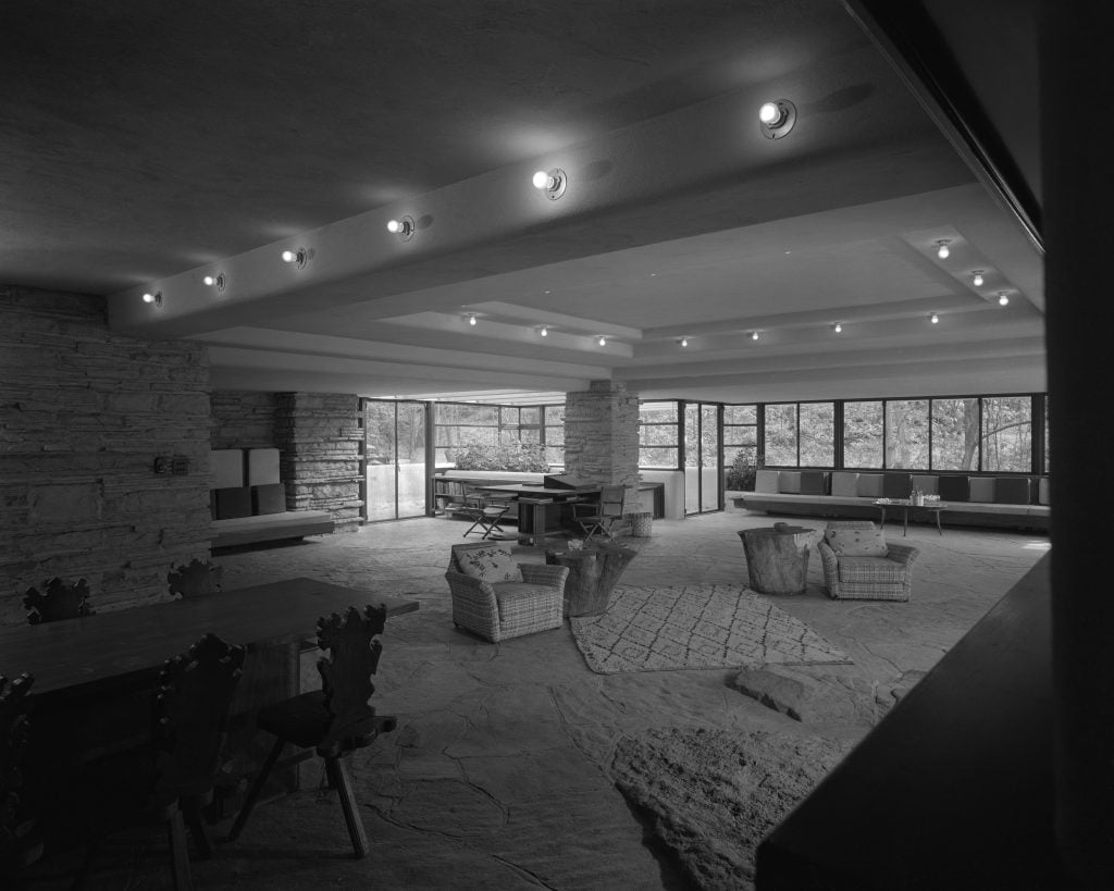 The interior of a home showing an open carpeted living room, sofa by a window, desk against a stone pillar, and armchairs dotted throughout the space