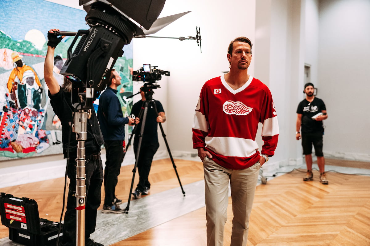 "Darren McCarty's Day Off" filming at the Detroit Institute of Arts on September 16, 2024. A man in a Red Wings jersey is in an art gallery, with cameras and crew around him. Danny DeKeyser Quilting Time (1986) by Romare Bearden.