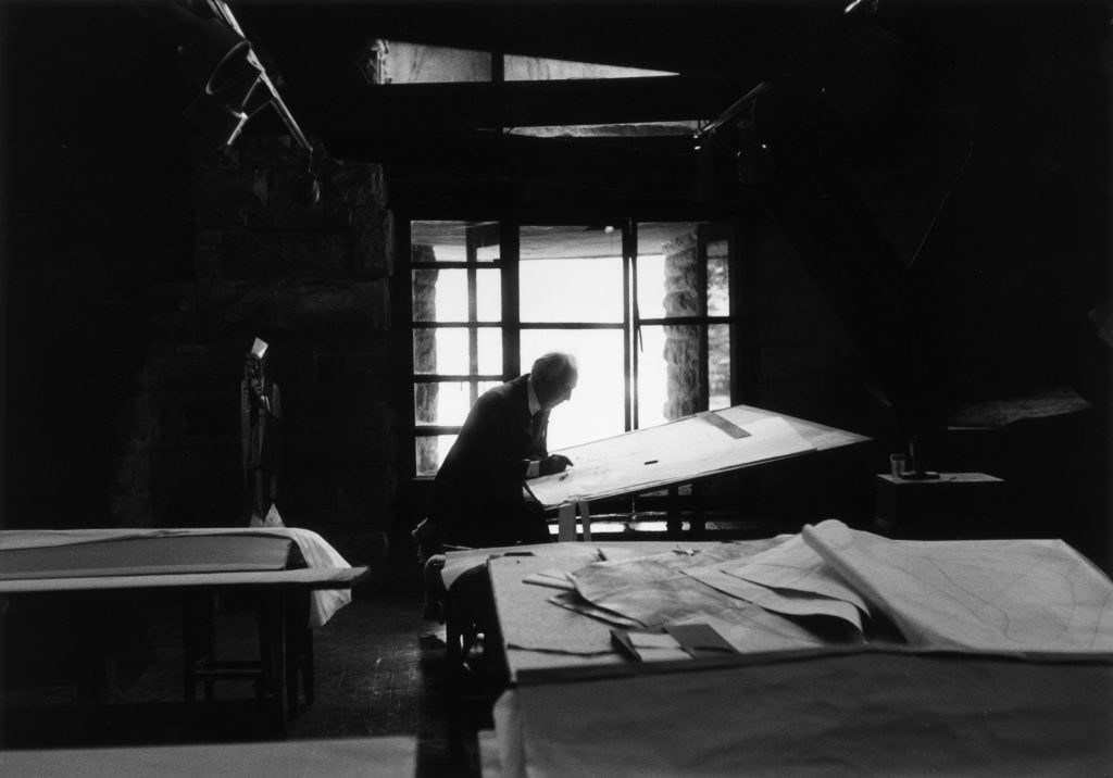 A man sitting at a drafting table is silhouetted against a bright glass door