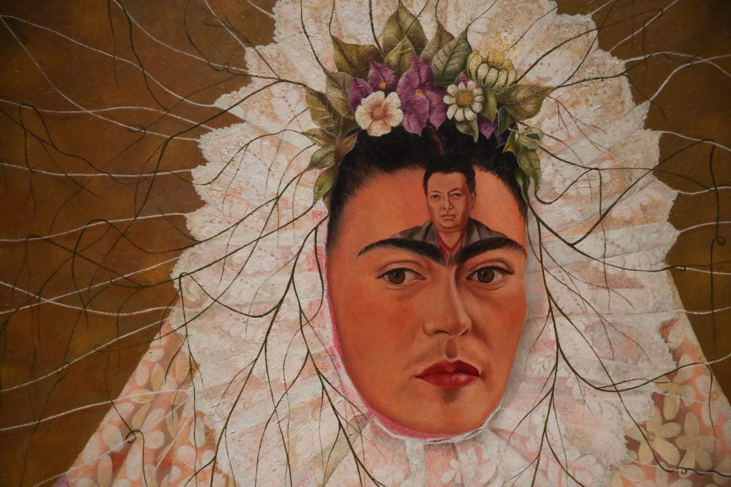 a painting of the artist Frida Kahlo dressed in tradiional tehuana mexican attired, with a white and lacy head piece and flowers. The painting of the artist show her husband, Diego Rivera, on her forehead.