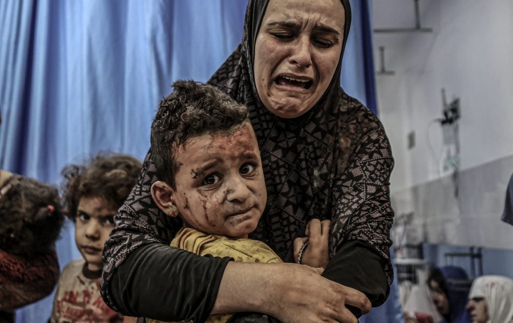 A sobbing mom holds her injured child in Al-Shifa Hospital in Gaza City after an Israeli airstrike on October 18, 2023. The boy is covered with blood and debris from the bombing, and looks terrified.