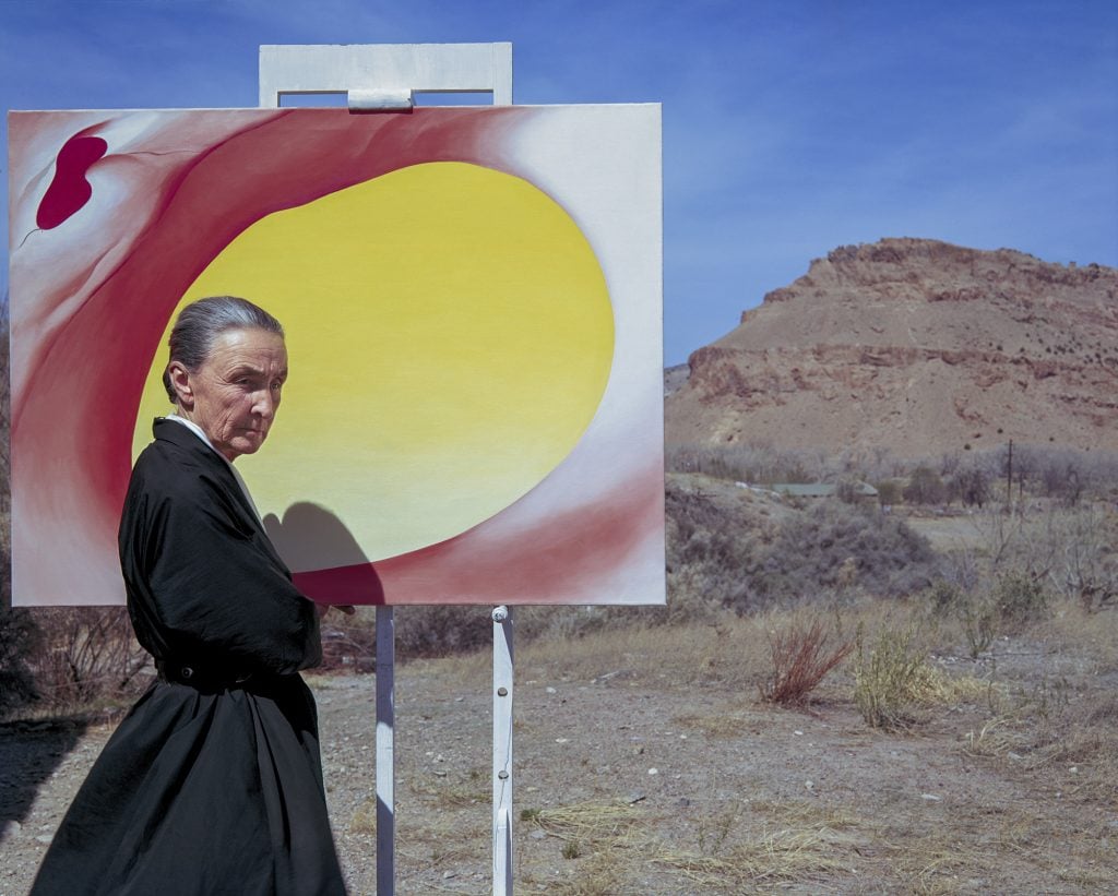 Artist Georgia O'Keeffe standing against a desert landscape, beside a large painting of an abstract yellow and red form