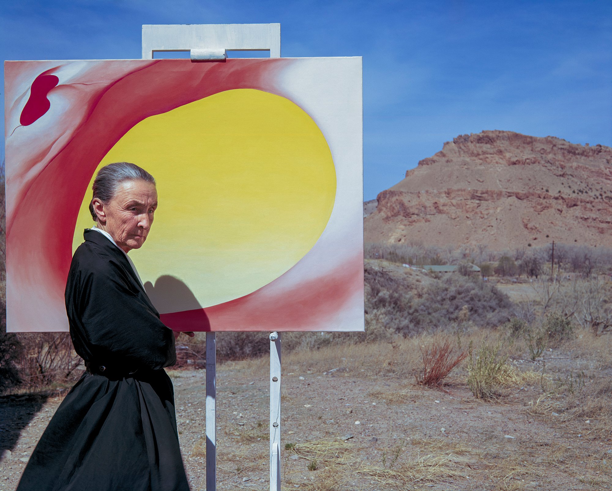 Artist Georgia O'Keeffe standing against a desert landscape, beside a large painting of an abstract yellow and red form