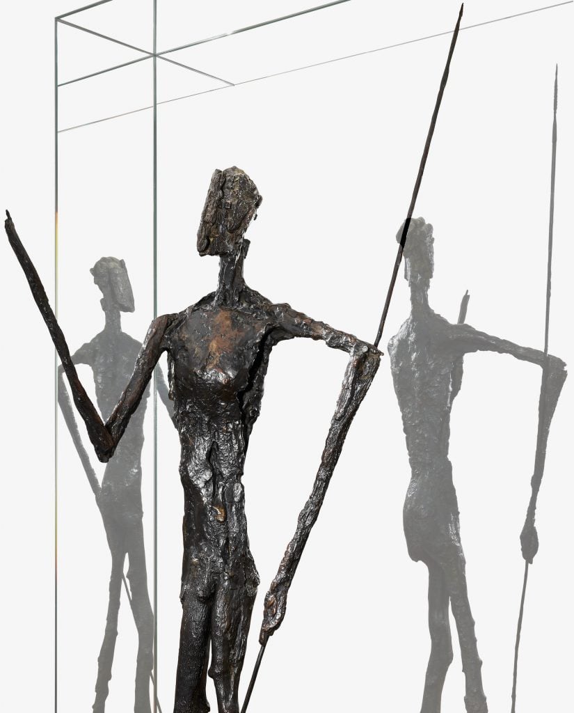 A detail of a bronze figurative sculpture with brown patina of an person standing holding a long spear-like stick, shown from the knees up with two reflections superimposed behind it.