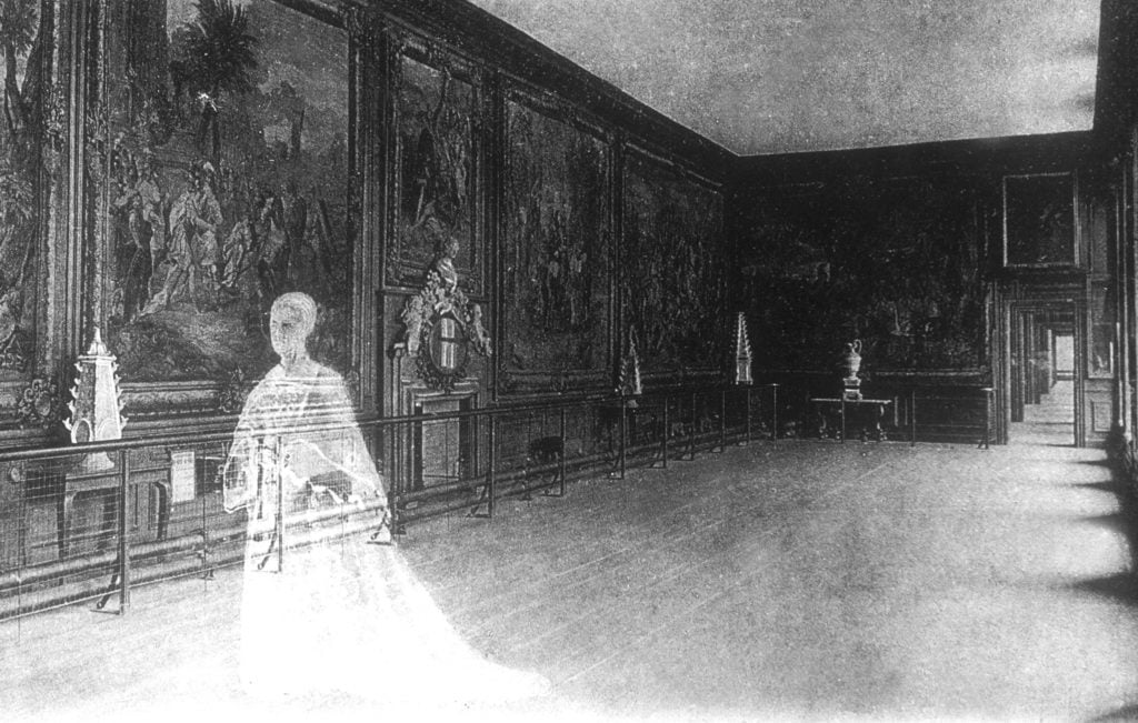 a faded old black and white photograph of a stately interior with art on the walls, the transparent, ghostly white figure of a woman in old-fashioned formal dress can be seen transposed over the interior scene to the left