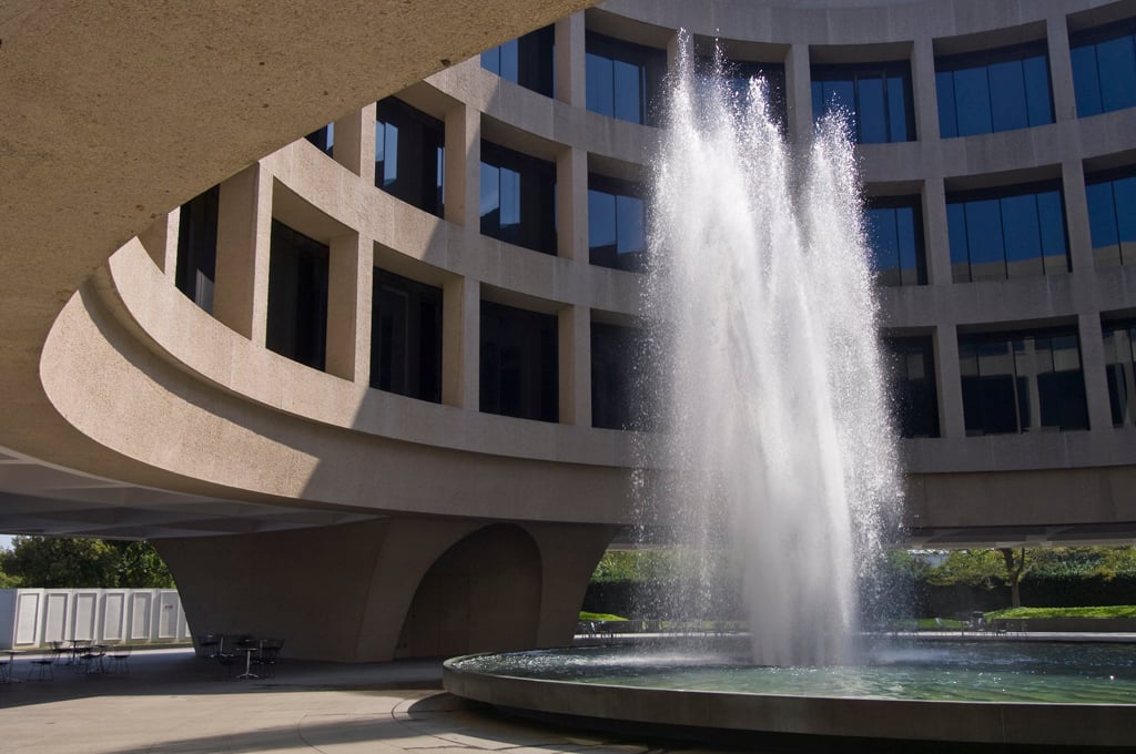 A large, circular fountain sprays water upward in an open courtyard surrounded by a curved concrete building with tall windows.