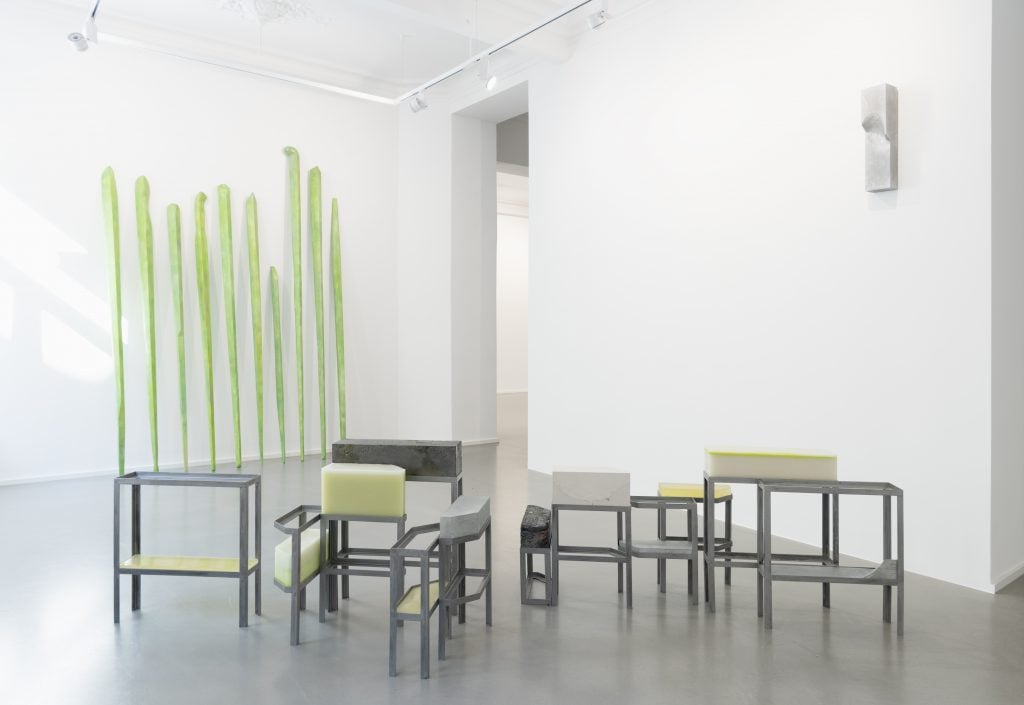 A series of metal table-like structures with beige blocks on them in a white gallery space; against the back wall a series of pale green sticks of various heights are leaned against the wall. All pieces by Gereon Krebber, and on view at Galerie Christian Lethert.