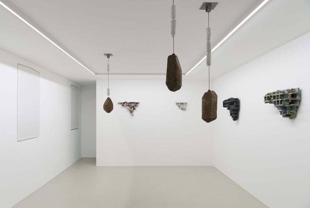 A white, windowless gallery space with glazed ceramic pieces with three-dimensional grids installed on the walls, and three variously placed dark objects hanging down from the ceiling, by artist Gereon Krebber and on view at Galerie Christian Lethert.