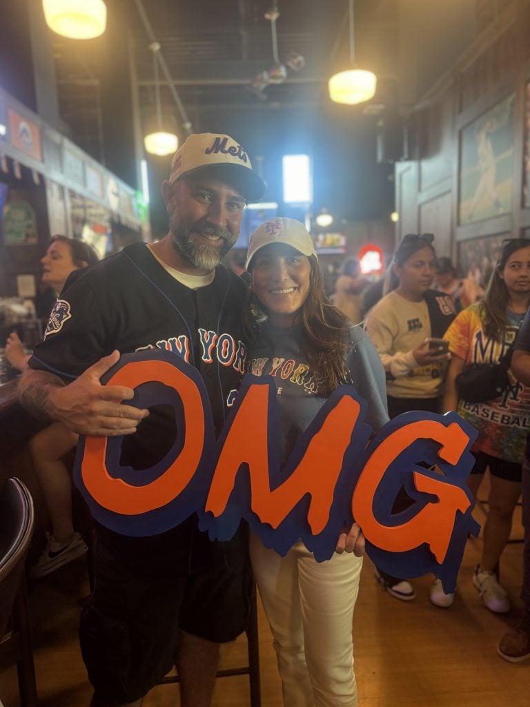 Artist Jerome McCroy with his OMG signs and Alex Cohen, wife of Mets owner Steve Cohen. They are in a crowded bar, wearing Mets shirts and hats. 