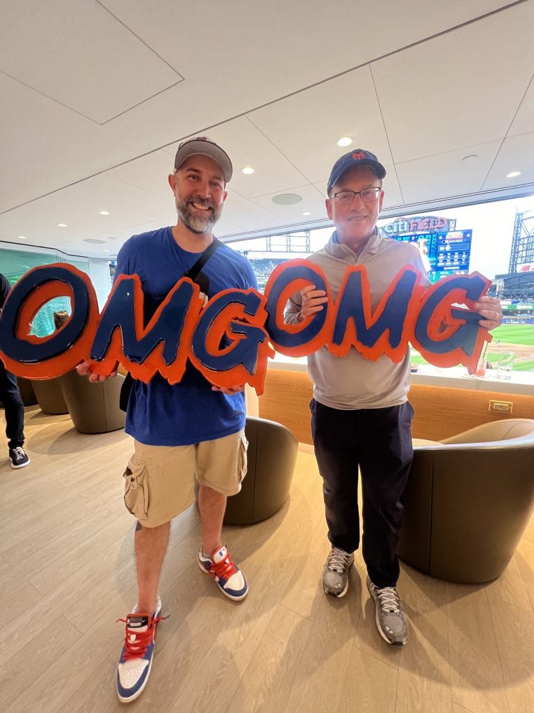 Artist Jerome McCroy with his OMG signs and Mets owner—and art collector—Steve Cohen. The two are in the owner's box at Citi Field, with a view out to home plate behind them. 