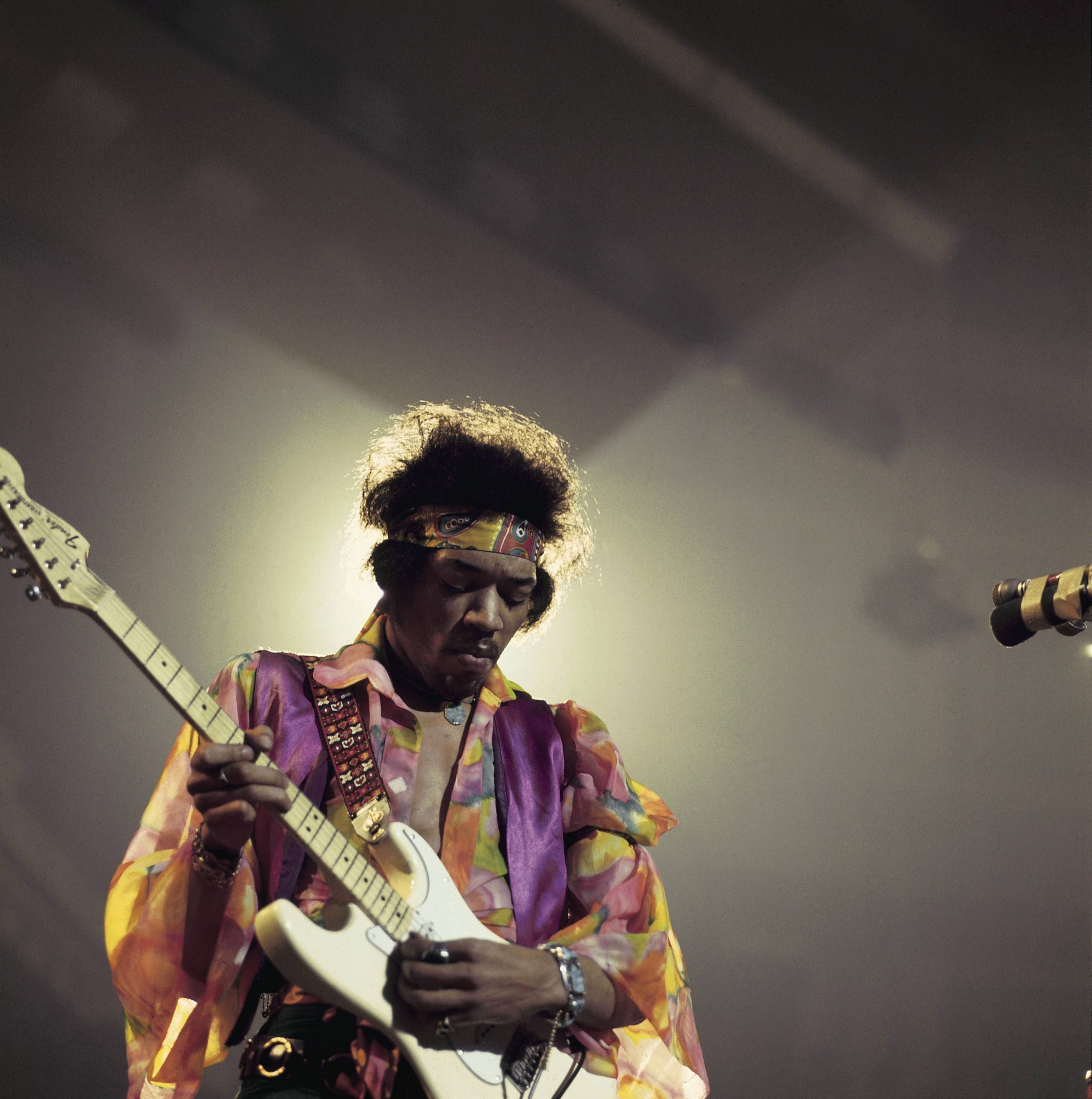 Musician Jimi Hendrix in a colorful top performs live on stage playing a white Fender Stratocaster guitar