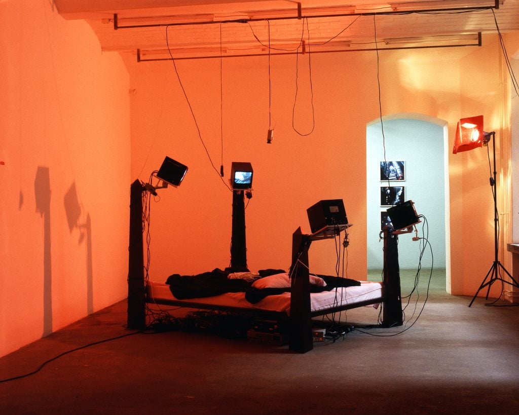 A bedframe by artist Julia Scher with CCTV monitors on all four corners and a stage light beside it making the room glow orange.