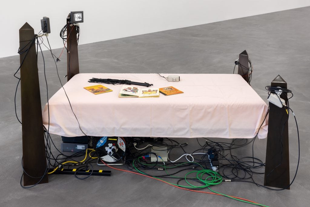 A sculpture by Agnes Scher of a bed with cameras and CCTV monitors on the top two corners, and an assortment of small books on top, made with a pink bedsheet.