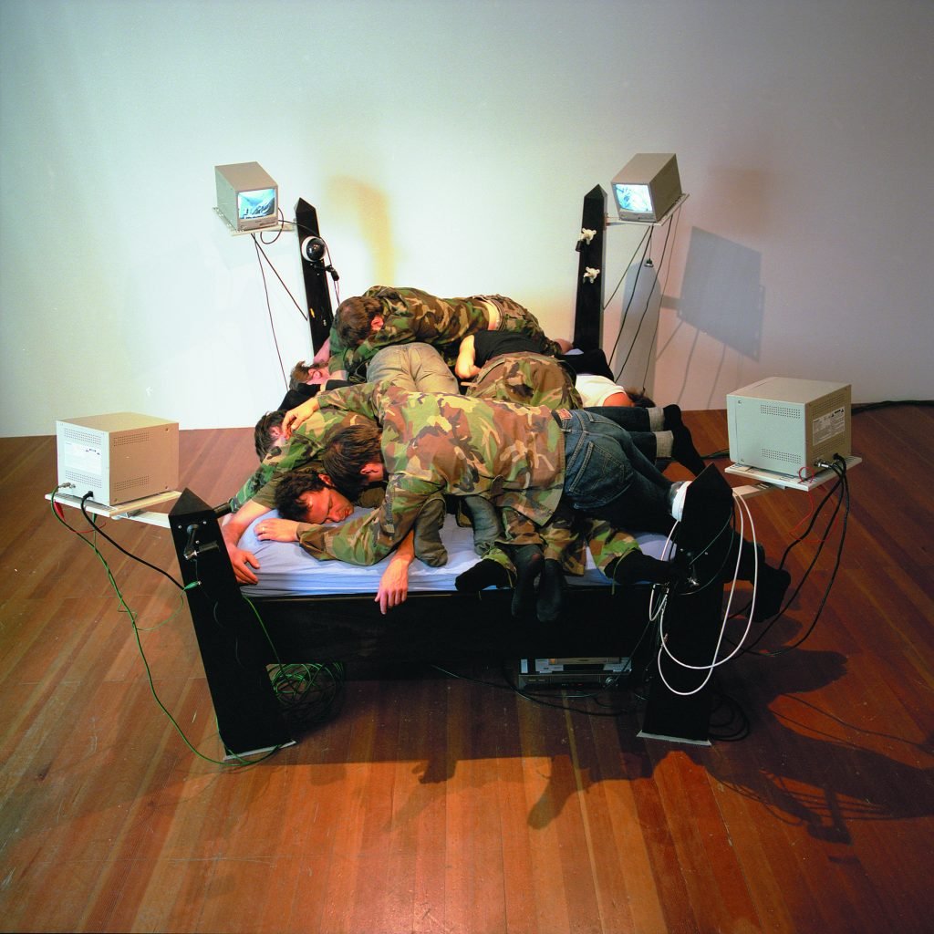 A bed with cameras and CCTV monitors on the four corners and a pile of men wearing camo lying on it, work by Agnes Scher.