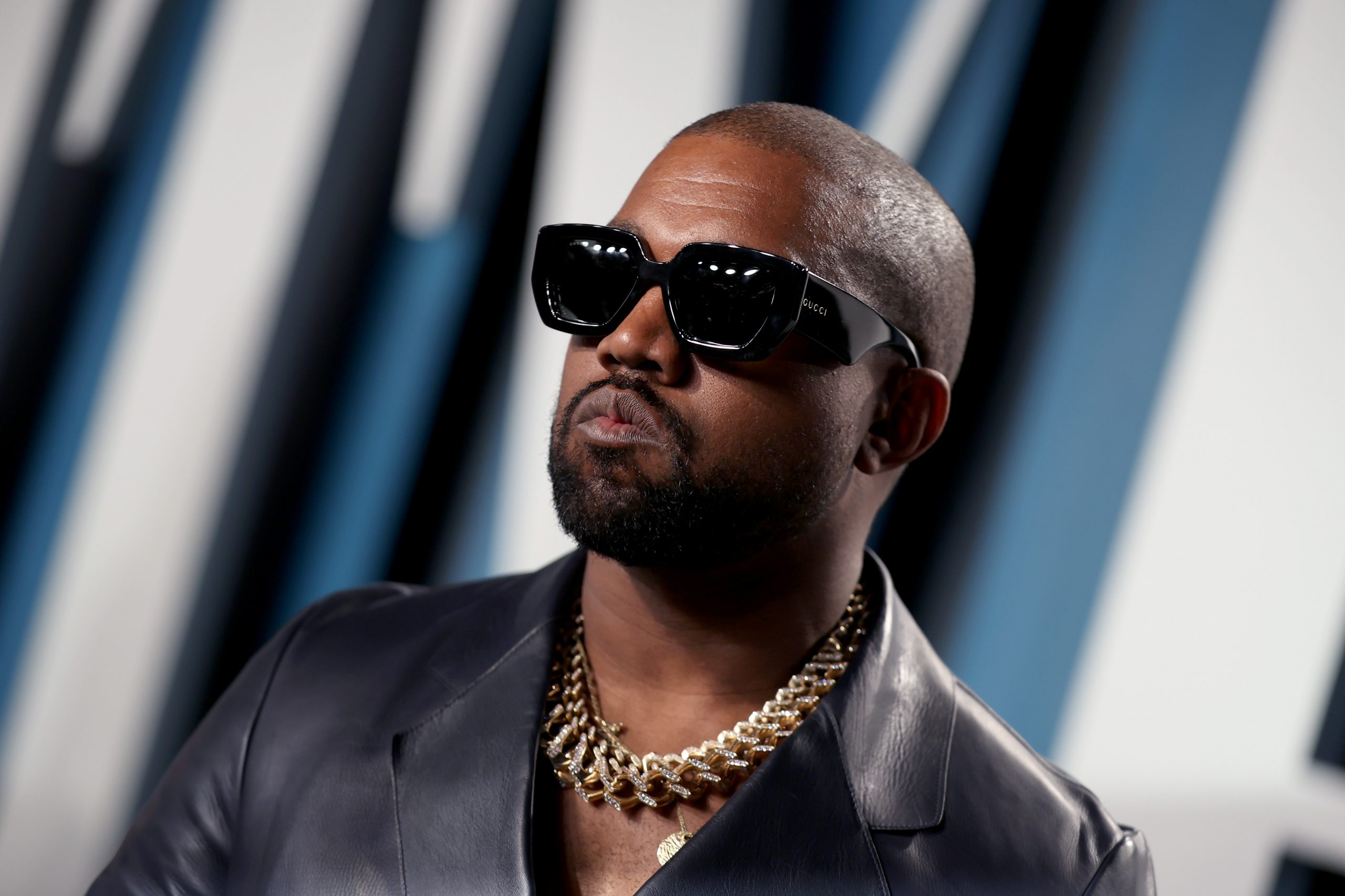 Kanye West wearing dark glasses, a gold chain, and a leather blazer