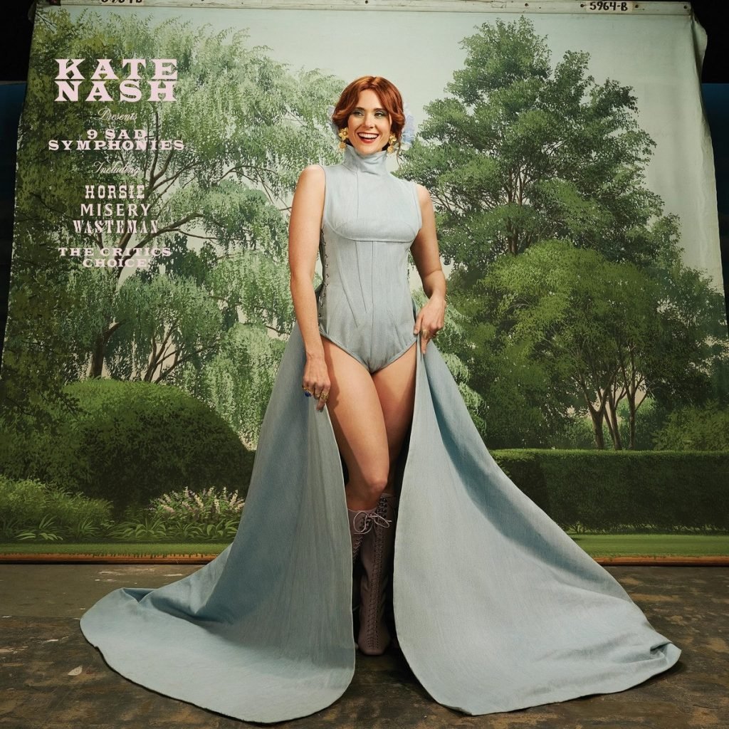 The cover of an album by Kate Nash showing her standing in a body suit with a large train, against a painted backdrop of trees