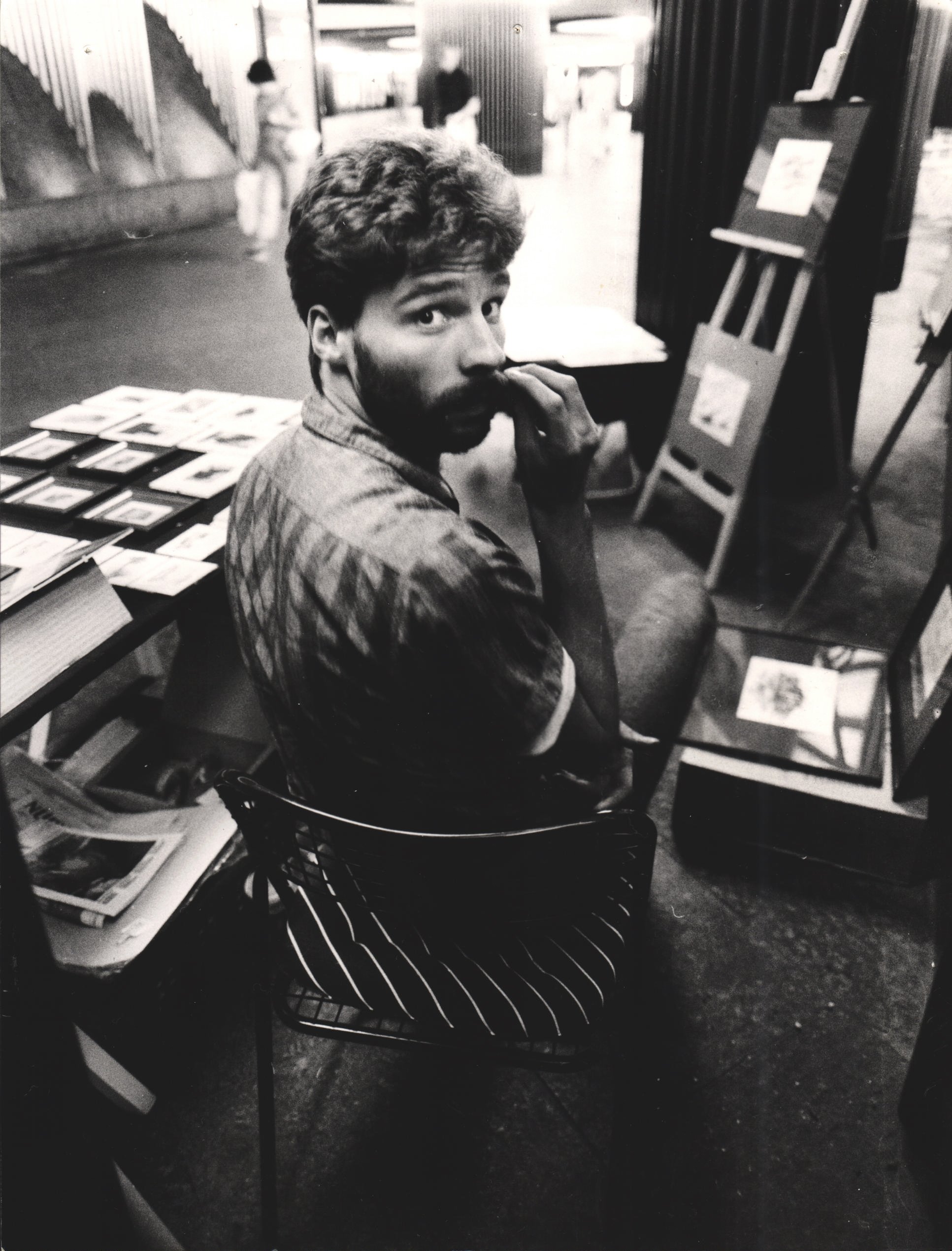 Black and white photo of Klaus Bode of Bode Galerie at a table selling prints in a subway passage looking back over his shoulder into the camera.