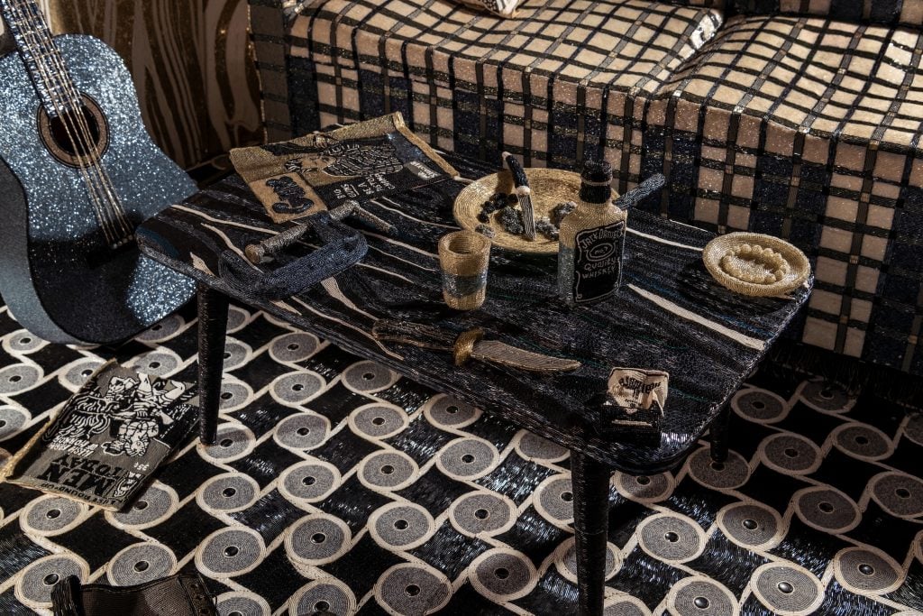 The interior of a mobile trailer where every object is covered in black and white beads. A coffee table on a linoleum floor, a whisky bottle, a hunting knife