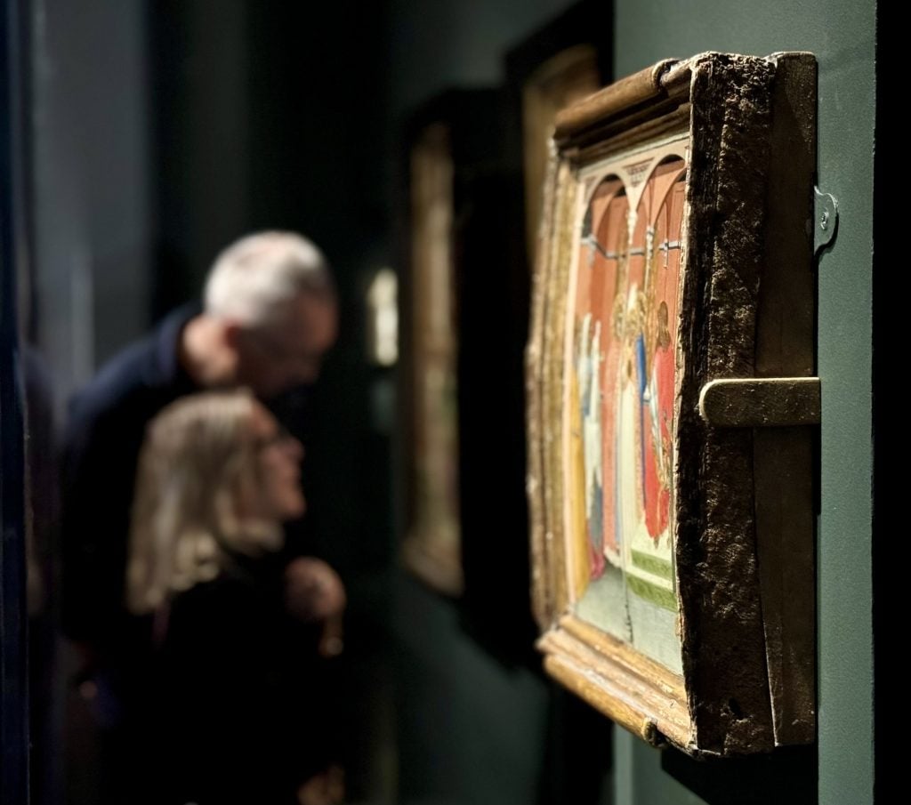 A small painting is seen from the side, as a couple look at another painting in the background