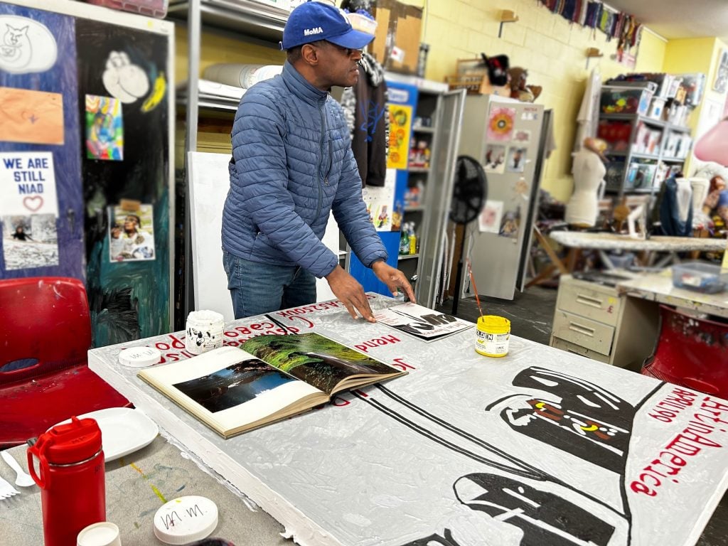 A Black man in an art studio, standing over a large artwork on a table