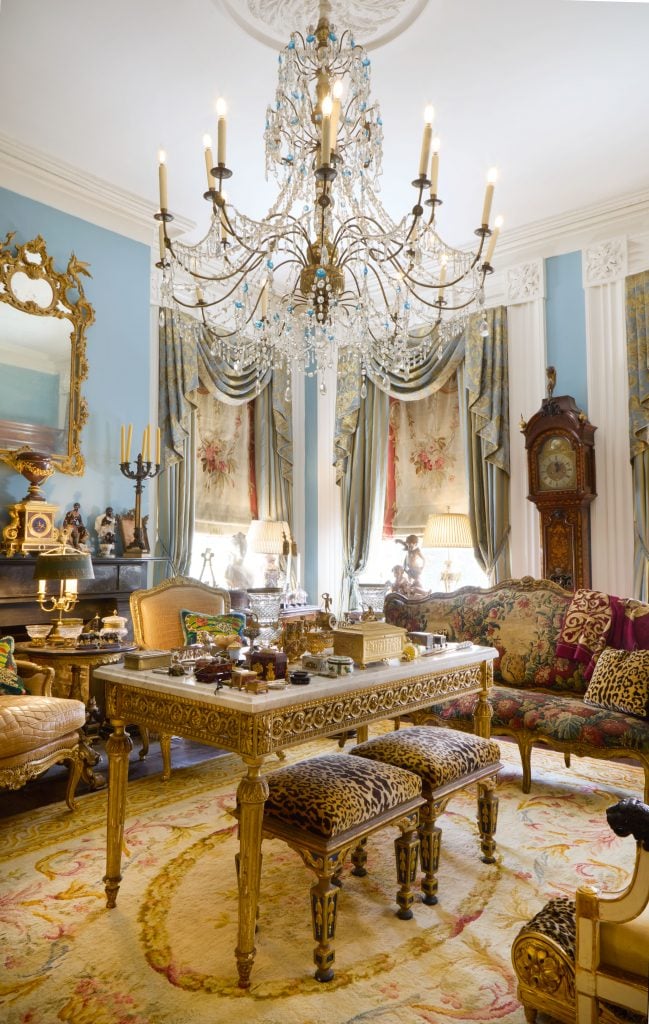 Inside an ornately decorated room with powder blue walls featuring items from the Kevin Stone and Mark Diamond Collection, including a crystal chandelier, gilt mirror, grandfather clock, and a rococo table covered in art objects.