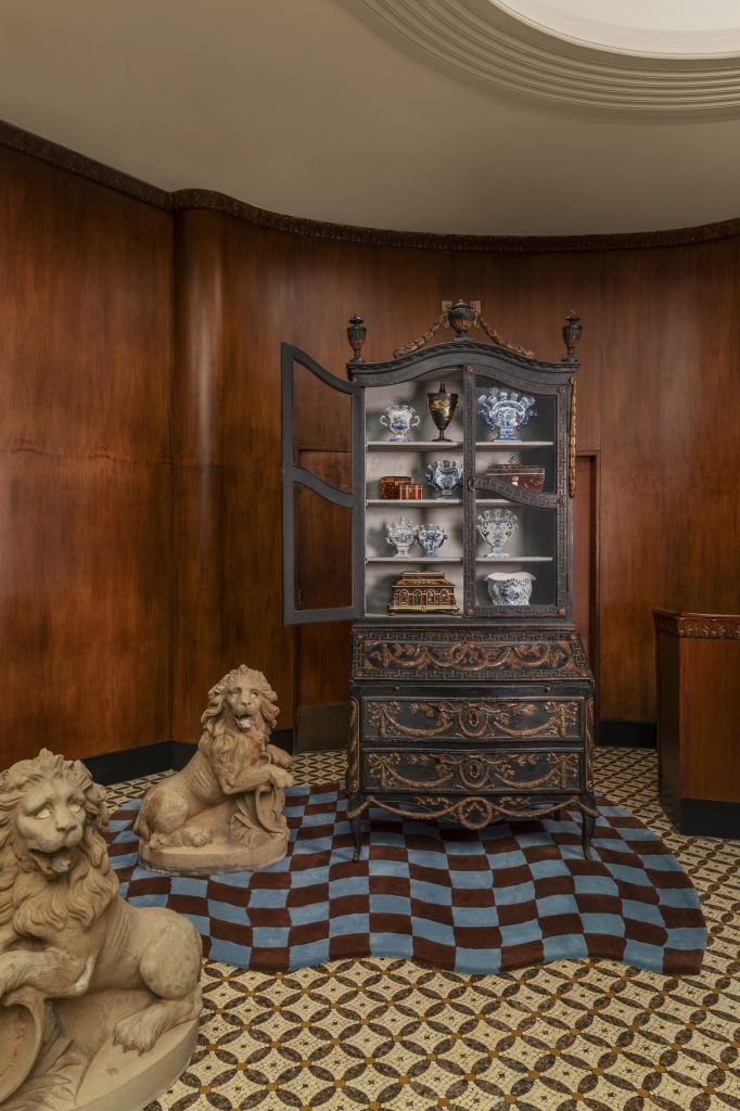 A wood-paneled gallery space at New Orleans Auction Galleries with a blue and brown checkered carpet over a yellow and gold crosshatch carpet, two stone lions and a wood chest of drawers with a display cabinet with an assortment of porcelain objects.