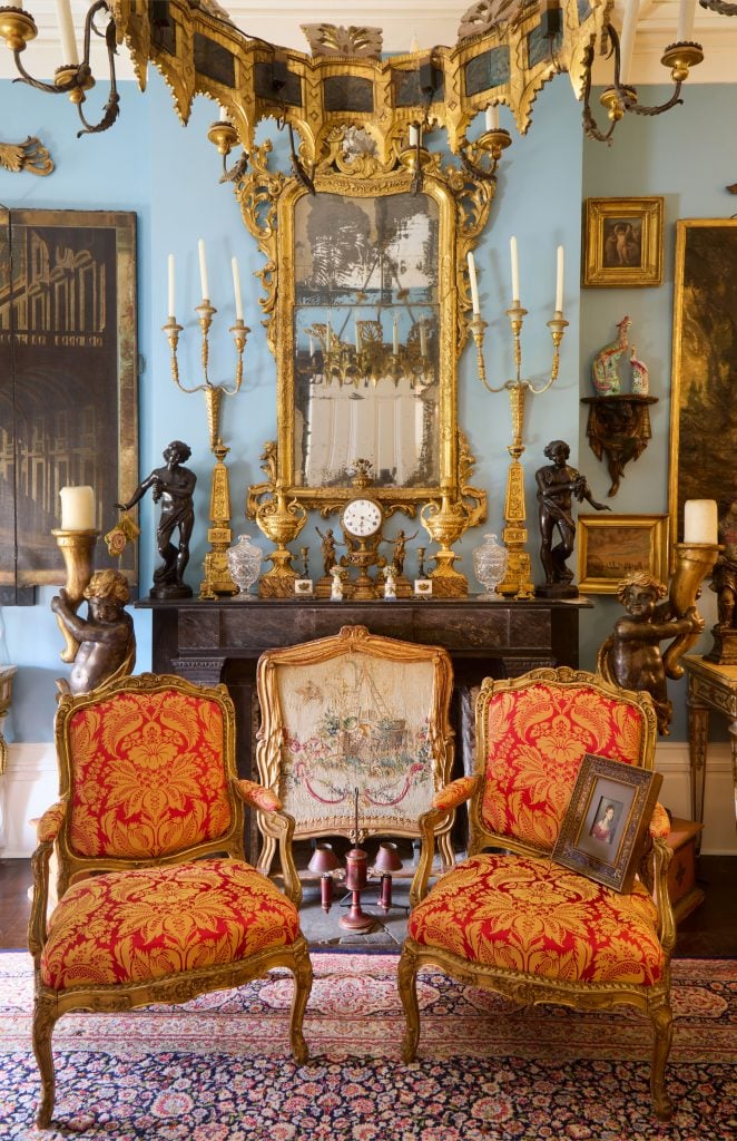 Interior view of an opulent room from the kevin stone and mark diamond collection showing two ornate upholstered chairs, a chandelier, gold framed mirror above a marble fireplace decorated with gilt objects.